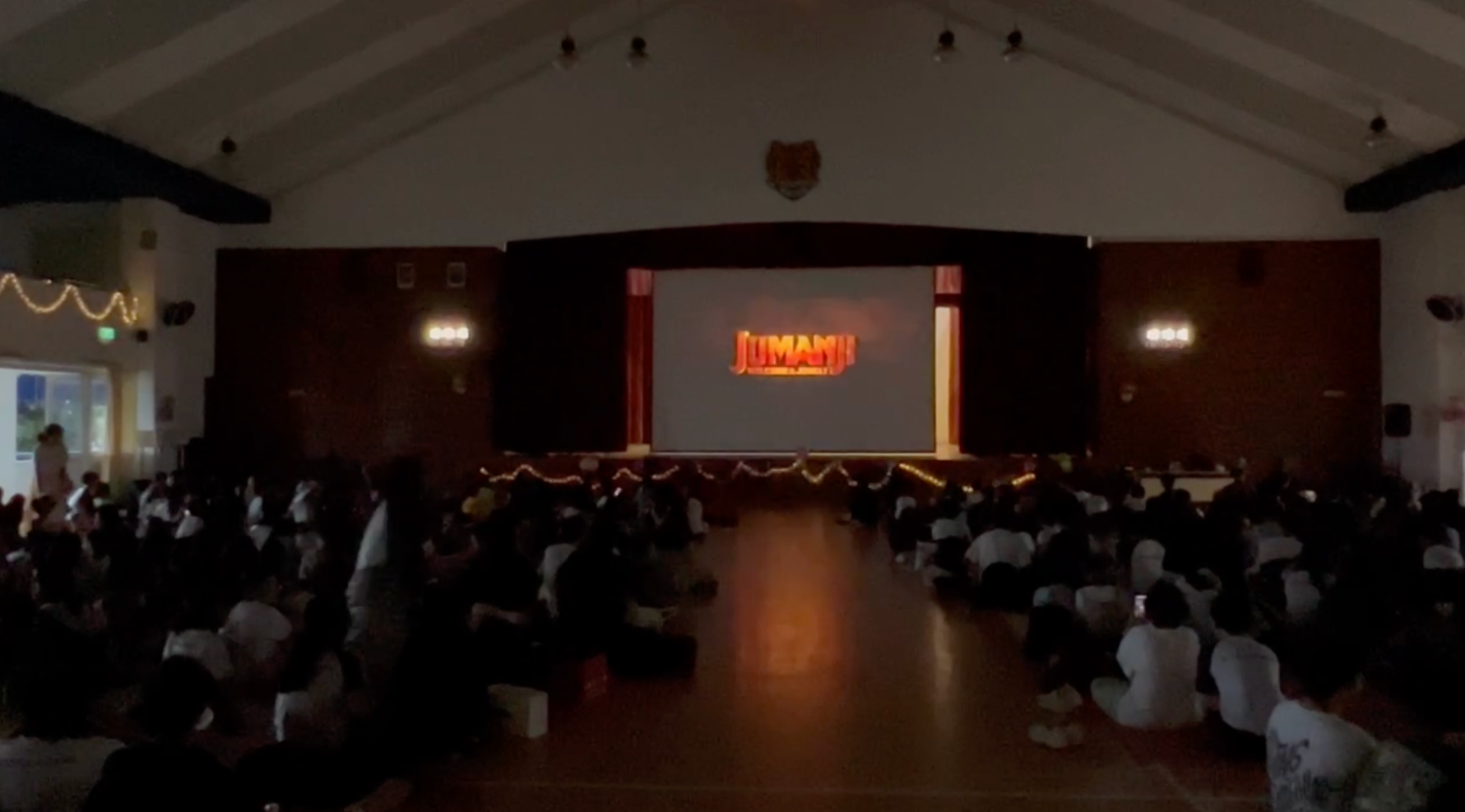 A large crowd of children sit on a hall floor watching the Jumanji title on a projected screen.